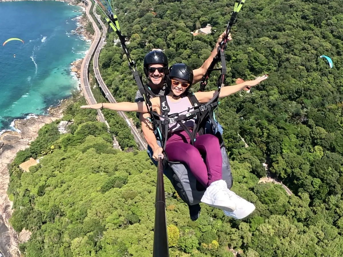 Passageira de braços abertos em voo de parapente sobre a floresta e a Avenida Niemeyer, com o mar ao fundo