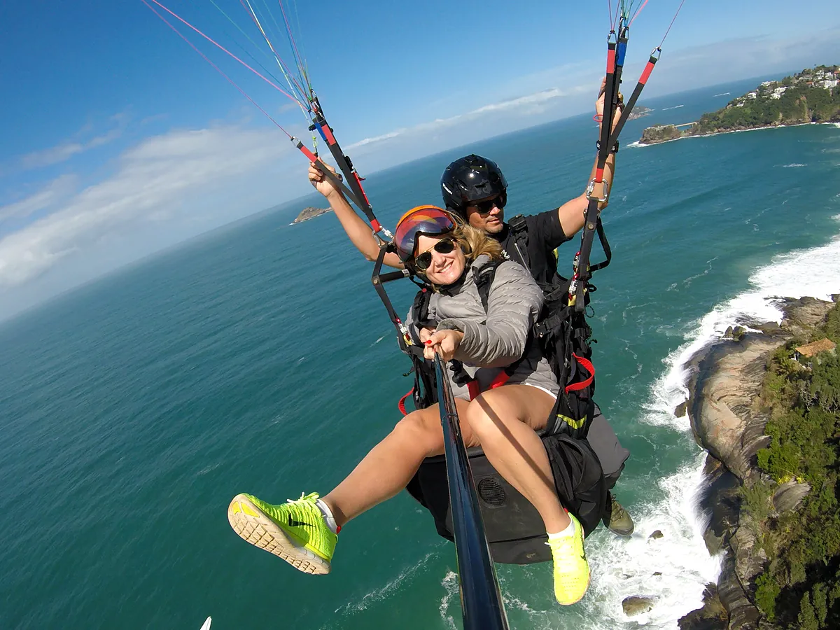 Passageira de braços abertos em voo duplo de parapente sobre o mar e o costão de São Conrado