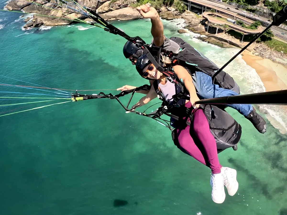 Passageira fazendo curva em voo duplo de parapente sobre o mar verde de São Conrado