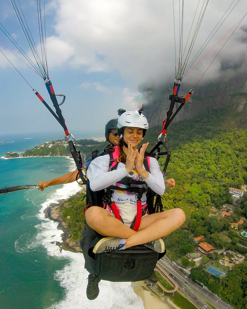 Passageira de pernas cruzadas e mãos unidas em voo de parapente sobre a Praia de São Conrado